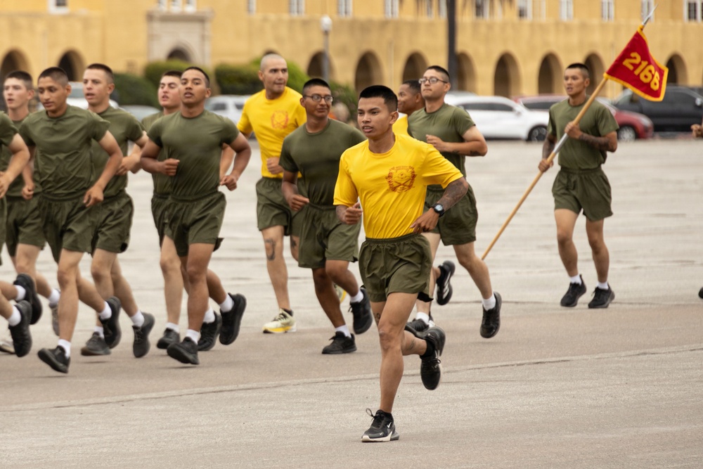 A formation of military recruits or trainees in physical training (PT) uniforms — olive green shorts and t-shirts — are running in formation on a parade deck or drill field. Two individuals wearing yellow t-shirts appear to be in leadership or instructor roles, with one carrying a guidon flag marked '2168'. The setting appears to be a military base, with a large yellow colonial-style building visible in the background.