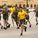 A formation of military recruits or trainees in physical training (PT) uniforms — olive green shorts and t-shirts — are running in formation on a parade deck or drill field. Two individuals wearing yellow t-shirts appear to be in leadership or instructor roles, with one carrying a guidon flag marked '2168'. The setting appears to be a military base, with a large yellow colonial-style building visible in the background.