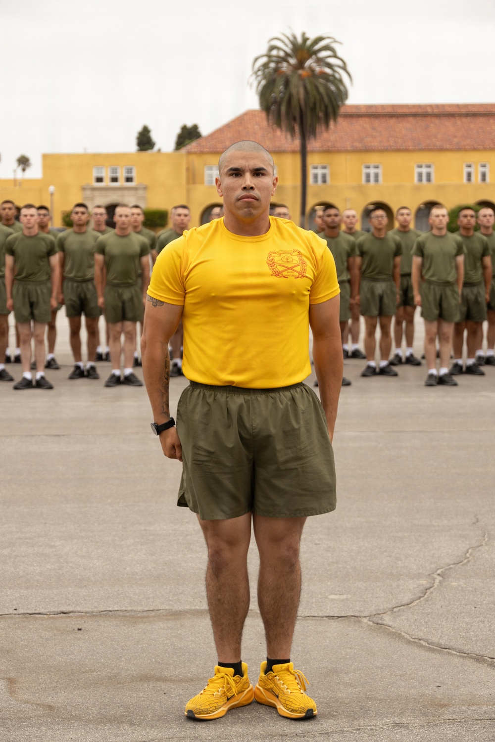 A military instructor or drill instructor stands at attention in the foreground wearing a yellow PT (physical training) shirt with an insignia, olive green shorts, and yellow athletic shoes. Behind him, multiple rows of uniformed military recruits or trainees stand at parade rest in matching olive green PT uniforms on a paved drill deck. The setting appears to be a military installation, likely Marine Corps Recruit Depot (MCRD) San Diego, based on the distinctive yellow Spanish Colonial Revival architecture visible in the background.