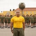 A military instructor or drill instructor stands at attention in the foreground wearing a yellow PT (physical training) shirt with an insignia, olive green shorts, and yellow athletic shoes. Behind him, multiple rows of uniformed military recruits or trainees stand at parade rest in matching olive green PT uniforms on a paved drill deck. The setting appears to be a military installation, likely Marine Corps Recruit Depot (MCRD) San Diego, based on the distinctive yellow Spanish Colonial Revival architecture visible in the background.