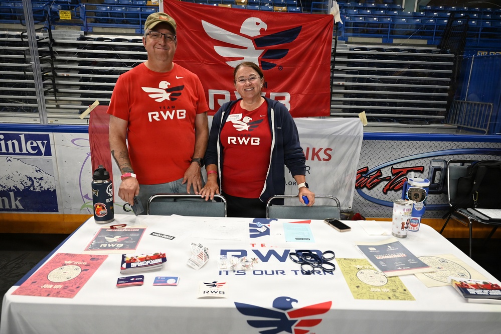 Two volunteers are staffing a Team RWB (Red, White &amp; Blue) information booth at what appears to be an indoor arena or sports facility. Both individuals are wearing matching red Team RWB shirts and standing behind a table covered with promotional materials, brochures, and merchandise. Team RWB is a nonprofit organization supporting military veterans through physical and social activity.