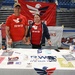 Two volunteers are staffing a Team RWB (Red, White &amp; Blue) information booth at what appears to be an indoor arena or sports facility. Both individuals are wearing matching red Team RWB shirts and standing behind a table covered with promotional materials, brochures, and merchandise. Team RWB is a nonprofit organization supporting military veterans through physical and social activity.