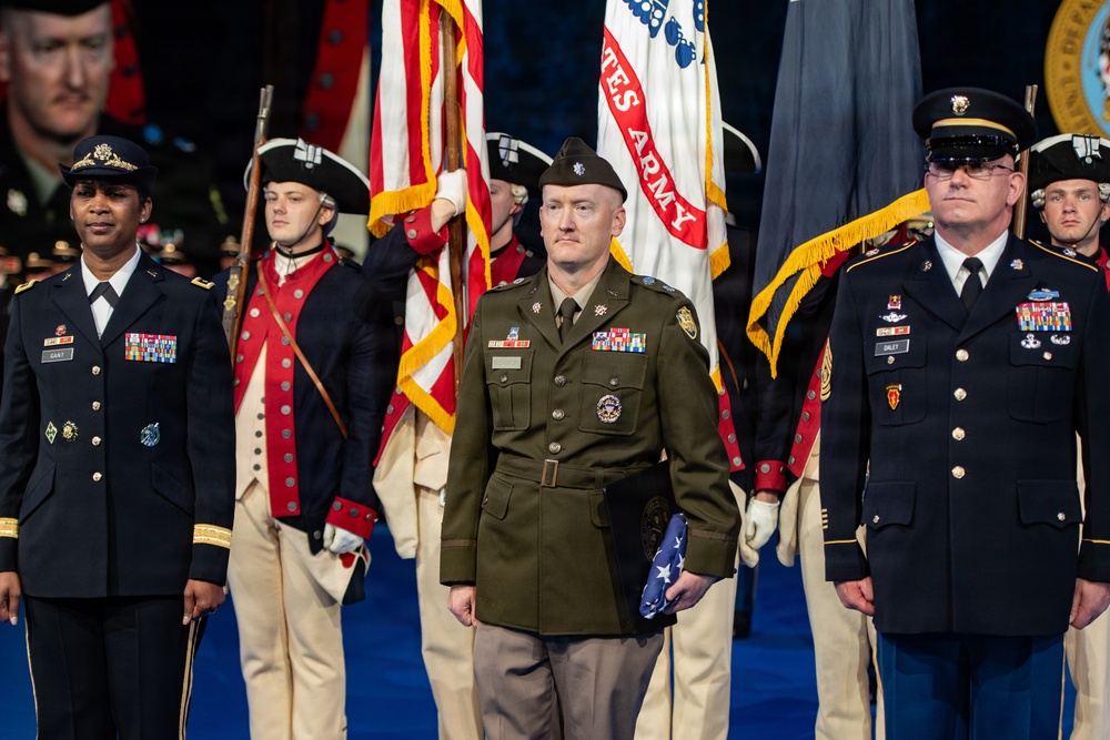 A formal U.S. Army military ceremony featuring personnel in multiple uniform types, including modern dress uniforms and historical Revolutionary War-era ceremonial uniforms. A central figure in an olive green Army Service Uniform holds a folded American flag, flanked by soldiers in blue dress uniforms. A color guard bearing U.S. and U.S. Army flags stands in the background.