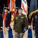 A formal U.S. Army military ceremony featuring personnel in multiple uniform types, including modern dress uniforms and historical Revolutionary War-era ceremonial uniforms. A central figure in an olive green Army Service Uniform holds a folded American flag, flanked by soldiers in blue dress uniforms. A color guard bearing U.S. and U.S. Army flags stands in the background.
