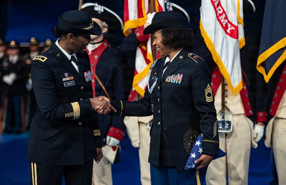 Two U.S. Army female soldiers are shaking hands during what appears to be a formal military ceremony. The officer on the left, identified by a name tape reading 'GANT,' appears to be a general officer based on visible rank insignia, while the soldier on the right holds a folded American flag and wears a distinctive black campaign hat. Color guard members and military flags, including one marked 'ARMY,' are visible in the background.
