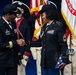 Two U.S. Army female soldiers are shaking hands during what appears to be a formal military ceremony. The officer on the left, identified by a name tape reading 'GANT,' appears to be a general officer based on visible rank insignia, while the soldier on the right holds a folded American flag and wears a distinctive black campaign hat. Color guard members and military flags, including one marked 'ARMY,' are visible in the background.