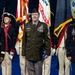 A formal U.S. Army military ceremony featuring three prominent figures standing at the forefront, with a color guard in historical Continental Army uniforms behind them. The central figure, wearing an olive green Army Service Uniform with a garrison cap, holds a folded American flag, suggesting a retirement or change-of-responsibility ceremony. The event appears to be held in a large indoor venue with Army insignia displayed in the background.