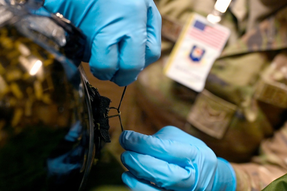 A person wearing blue nitrile gloves and military camouflage uniform is handling a small dark object, possibly a sample or material fragment. The individual is wearing a visible ID badge with a US flag and what appears to be a military or government insignia. The close-up shot suggests a careful, procedural handling of the item.