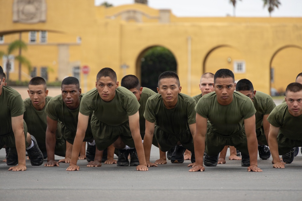 A large group of military recruits in olive green PT (physical training) uniforms are performing push-ups in unison on a paved surface. They are positioned in organized rows facing the camera, with a distinctive yellow military building visible in the background. The setting and uniforms are consistent with a U.S. Marine Corps recruit training depot.