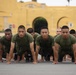 A large group of military recruits in olive green PT (physical training) uniforms are performing push-ups in unison on a paved surface. They are positioned in organized rows facing the camera, with a distinctive yellow military building visible in the background. The setting and uniforms are consistent with a U.S. Marine Corps recruit training depot.