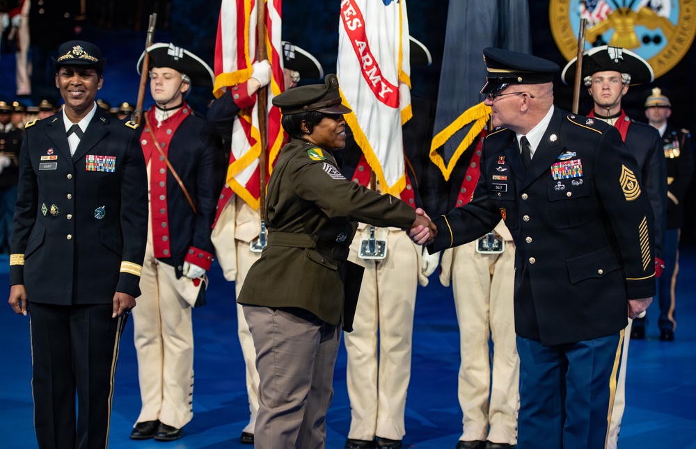 A formal U.S. Army military ceremony is taking place on a blue-floored stage or arena. Two soldiers in the foreground are shaking hands, while a female officer stands to the left observing. A color guard in historical Revolutionary War-era uniforms stands in the background holding flags including a United States Army flag.