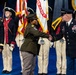 A formal U.S. Army military ceremony is taking place on a blue-floored stage or arena. Two soldiers in the foreground are shaking hands, while a female officer stands to the left observing. A color guard in historical Revolutionary War-era uniforms stands in the background holding flags including a United States Army flag.