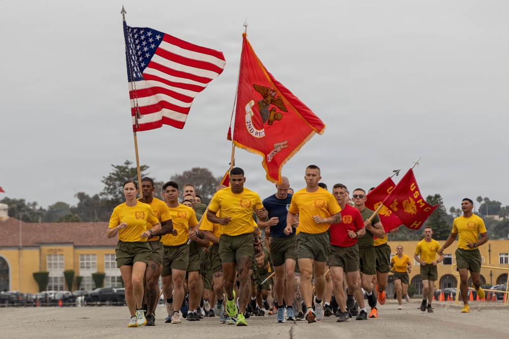 A large group of U.S. Marine Corps personnel are conducting a formation run, carrying the American flag and USMC unit guidons. The runners are dressed in standard PT (physical training) uniforms consisting of yellow t-shirts and olive/khaki shorts. The setting appears to be a military installation, likely MCRD (Marine Corps Recruit Depot) San Diego, based on the distinctive yellow buildings visible in the background.