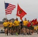 A large group of U.S. Marine Corps personnel are conducting a formation run, carrying the American flag and USMC unit guidons. The runners are dressed in standard PT (physical training) uniforms consisting of yellow t-shirts and olive/khaki shorts. The setting appears to be a military installation, likely MCRD (Marine Corps Recruit Depot) San Diego, based on the distinctive yellow buildings visible in the background.