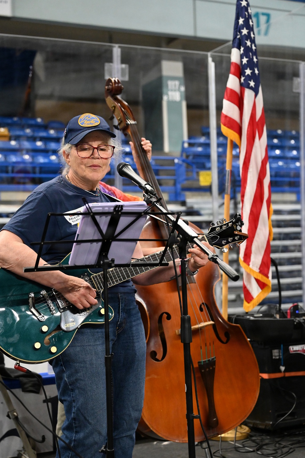 An older woman wearing a blue cap and red-framed glasses plays a green Gretsch electric guitar at what appears to be an indoor arena or sports venue. She stands at a music stand with sheet music and a microphone, while another musician plays an upright double bass behind her. An American flag is visible to the right, and empty blue stadium seats are seen in the background.