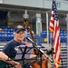 An older woman wearing a blue cap and red-framed glasses plays a green Gretsch electric guitar at what appears to be an indoor arena or sports venue. She stands at a music stand with sheet music and a microphone, while another musician plays an upright double bass behind her. An American flag is visible to the right, and empty blue stadium seats are seen in the background.