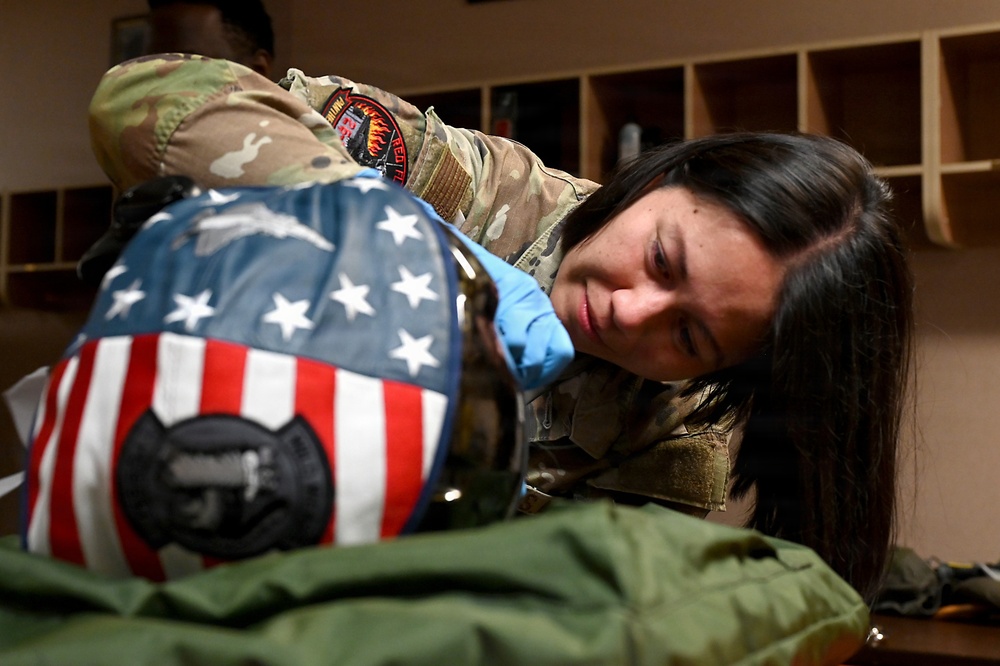 A female military service member in MultiCam/OCP camouflage uniform is closely inspecting or working on a flight helmet painted with an American flag design. She is wearing blue latex gloves and appears to be conducting a detailed examination or maintenance task. The setting appears to be an equipment room or locker area with wooden shelving visible in the background.
