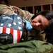 A female military service member in MultiCam/OCP camouflage uniform is closely inspecting or working on a flight helmet painted with an American flag design. She is wearing blue latex gloves and appears to be conducting a detailed examination or maintenance task. The setting appears to be an equipment room or locker area with wooden shelving visible in the background.