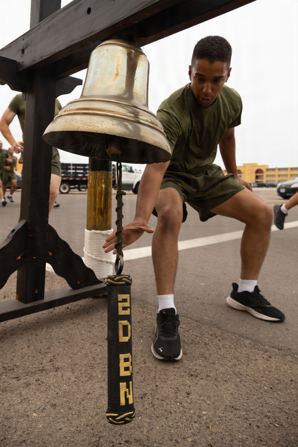 A young man in olive green military physical training attire reaches toward a brass bell mounted on a black wooden frame. The bell's pull rope is decorated with a black and gold lanyard labeled '2D BN' (2nd Battalion). Other individuals in similar PT gear are visible in the background on what appears to be a military installation.