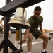 A young man in olive green military physical training attire reaches toward a brass bell mounted on a black wooden frame. The bell's pull rope is decorated with a black and gold lanyard labeled '2D BN' (2nd Battalion). Other individuals in similar PT gear are visible in the background on what appears to be a military installation.