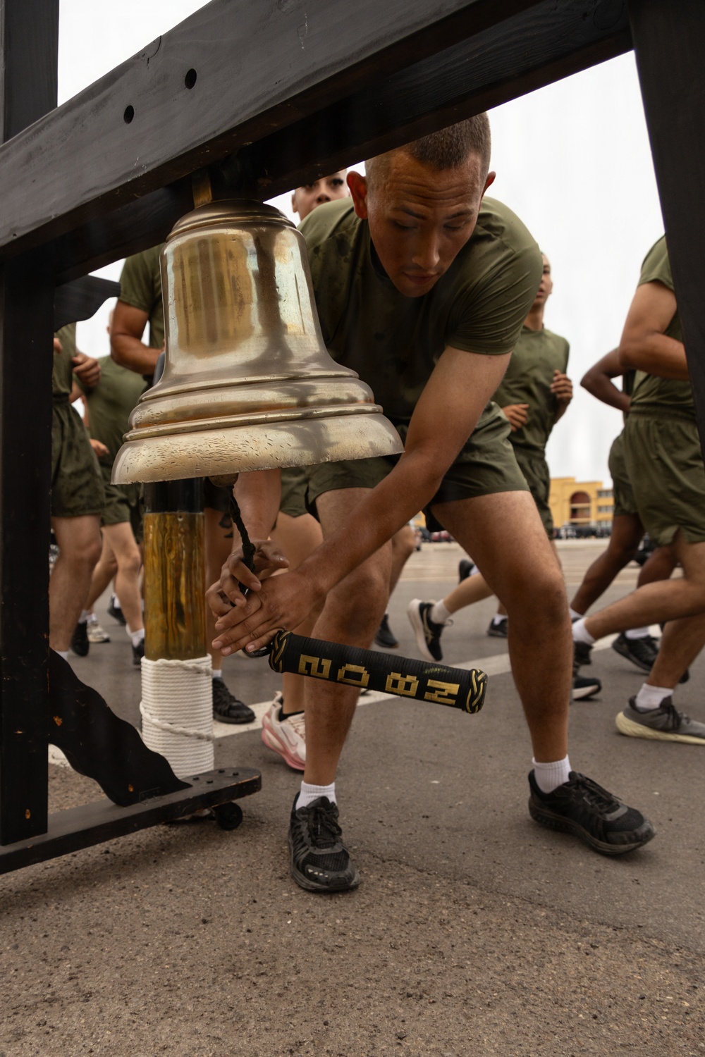 A military trainee in olive green PT (physical training) uniform is ringing a large brass bell mounted on a black wooden frame, using a mallet with black and gold grip tape. Other trainees in identical uniforms are running in the background on what appears to be a paved outdoor area. This appears to be a military training event, possibly a graduation or selection ceremony where ringing the bell carries symbolic significance.