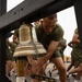 A military trainee in olive green PT (physical training) uniform is ringing a large brass bell mounted on a black wooden frame, using a mallet with black and gold grip tape. Other trainees in identical uniforms are running in the background on what appears to be a paved outdoor area. This appears to be a military training event, possibly a graduation or selection ceremony where ringing the bell carries symbolic significance.