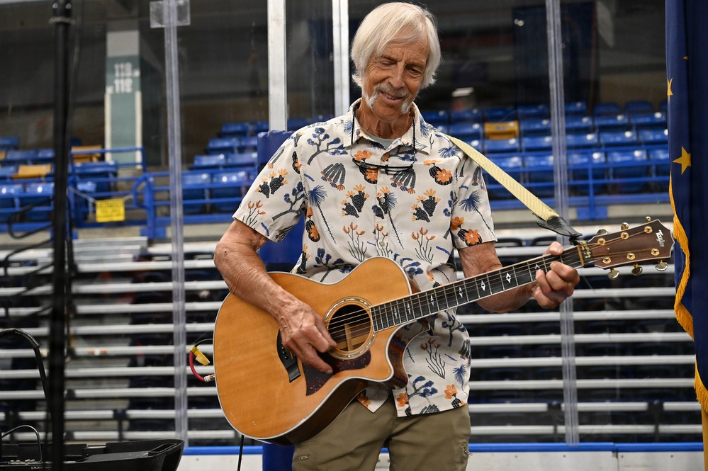 An elderly man with white hair is playing an acoustic guitar in what appears to be an indoor sports arena or ice rink. He is wearing a colorful floral/botanical print short-sleeve shirt and khaki shorts, smiling while performing. Blue stadium seating and a partial flag are visible in the background.