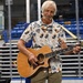 An elderly man with white hair is playing an acoustic guitar in what appears to be an indoor sports arena or ice rink. He is wearing a colorful floral/botanical print short-sleeve shirt and khaki shorts, smiling while performing. Blue stadium seating and a partial flag are visible in the background.