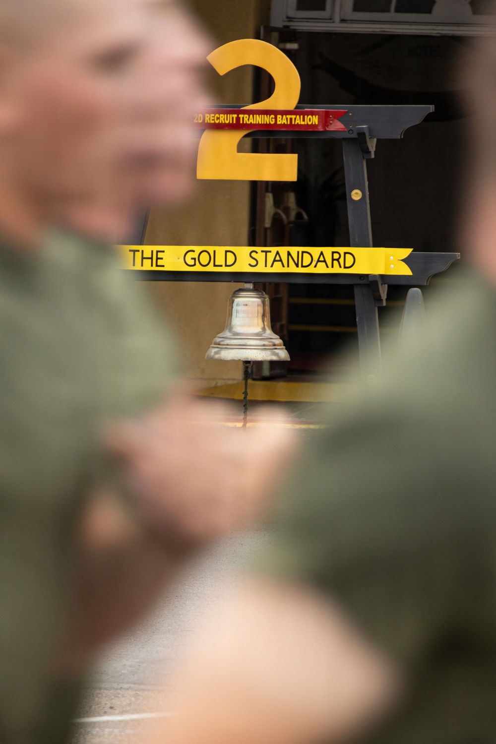 The image shows a military training environment associated with the 2nd Recruit Training Battalion, featuring a prominent display sign reading 'THE GOLD STANDARD' with a large yellow numeral '2' and a hanging metal bell. Blurred figures in olive/green military uniforms occupy the foreground. The setting appears to be a U.S. Marine Corps recruit training facility.