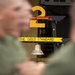 The image shows a military training environment associated with the 2nd Recruit Training Battalion, featuring a prominent display sign reading 'THE GOLD STANDARD' with a large yellow numeral '2' and a hanging metal bell. Blurred figures in olive/green military uniforms occupy the foreground. The setting appears to be a U.S. Marine Corps recruit training facility.