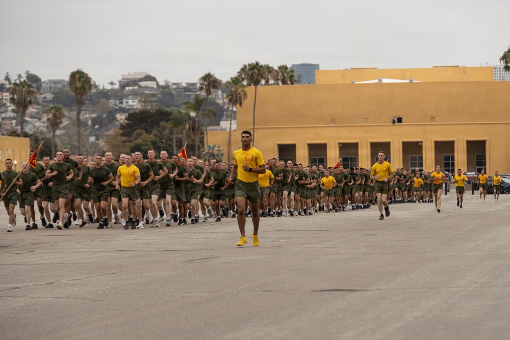 A large formation of military personnel, likely U.S. Marines, conducting a group physical training run on a paved parade deck or tarmac. The majority wear olive green t-shirts and shorts, while leaders at the front wear distinctive yellow shirts and shoes. The setting appears to be a military installation, consistent with Marine Corps Recruit Depot (MCRD) San Diego based on the architecture and surrounding landscape.