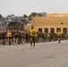 A large formation of military personnel, likely U.S. Marines, conducting a group physical training run on a paved parade deck or tarmac. The majority wear olive green t-shirts and shorts, while leaders at the front wear distinctive yellow shirts and shoes. The setting appears to be a military installation, consistent with Marine Corps Recruit Depot (MCRD) San Diego based on the architecture and surrounding landscape.