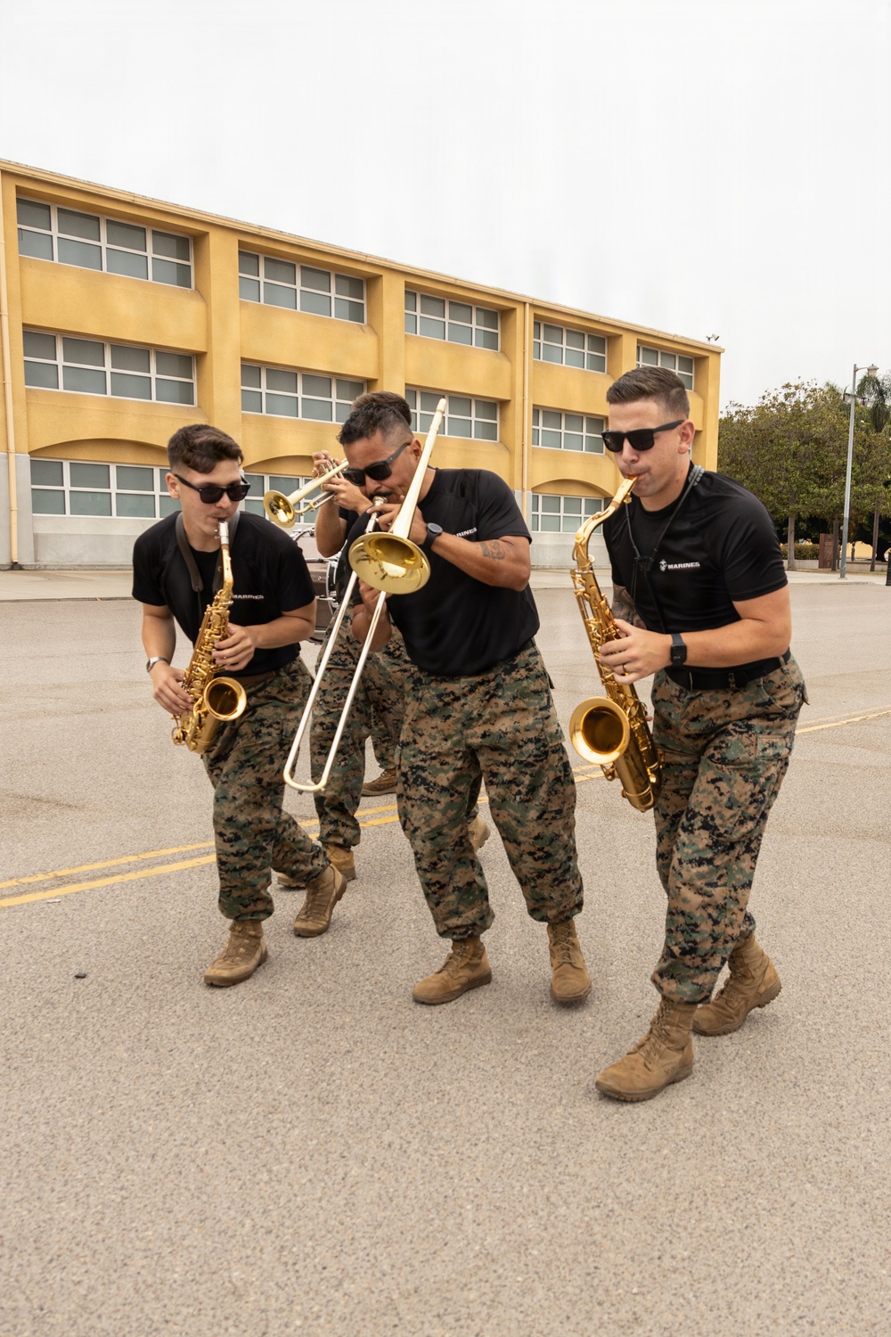 Three U.S. Marines in camouflage utility uniforms and black t-shirts are playing brass and woodwind instruments while walking across a paved outdoor area. The musicians are performing energetically, with two playing saxophones and one playing a trombone, with a trumpet player partially visible in the background. The setting appears to be a military installation, identifiable by the institutional building and the 'MARINES' text visible on their shirts.