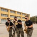 Three U.S. Marines in camouflage utility uniforms and black t-shirts are playing brass and woodwind instruments while walking across a paved outdoor area. The musicians are performing energetically, with two playing saxophones and one playing a trombone, with a trumpet player partially visible in the background. The setting appears to be a military installation, identifiable by the institutional building and the 'MARINES' text visible on their shirts.