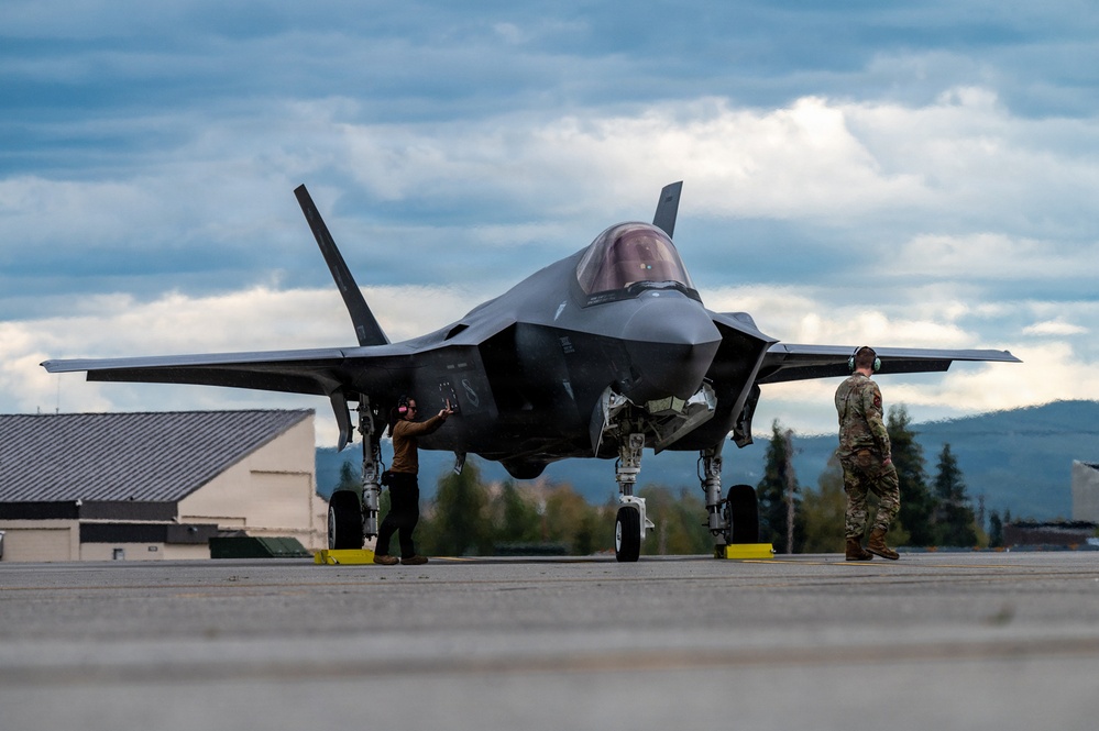 A fifth-generation stealth fighter jet, identifiable as an F-35 Lightning II, is parked on a flight line apron at what appears to be a military airbase. Two ground crew personnel wearing hearing protection are conducting post-flight or pre-flight servicing around the aircraft. The setting features mountainous terrain, coniferous trees, and overcast skies consistent with a northern or Alaskan environment.