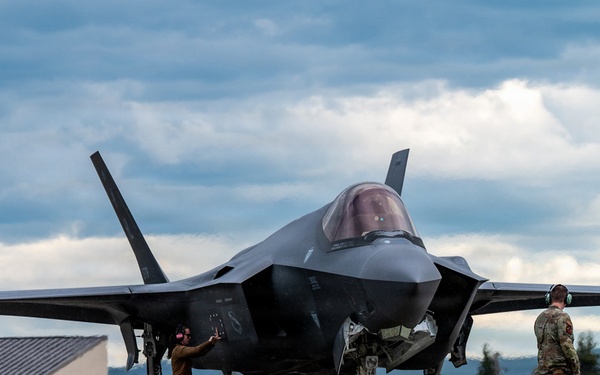 A fifth-generation stealth fighter jet, identifiable as an F-35 Lightning II, is parked on a flight line apron at what appears to be a military airbase. Two ground crew personnel wearing hearing protection are conducting post-flight or pre-flight servicing around the aircraft. The setting features mountainous terrain, coniferous trees, and overcast skies consistent with a northern or Alaskan environment.