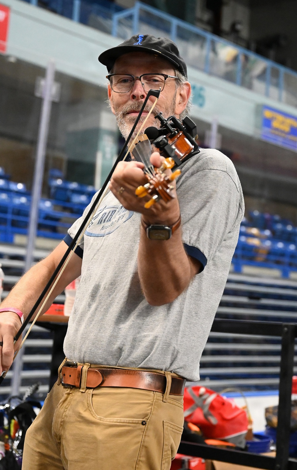 A middle-aged man with a beard and glasses is playing a fiddle/violin in what appears to be an indoor arena or sports facility. He is wearing a gray t-shirt, khaki pants, a brown leather belt, and a black baseball cap. A microphone or pickup device is attached to the instrument's scroll area.