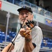 A middle-aged man with a beard and glasses is playing a fiddle/violin in what appears to be an indoor arena or sports facility. He is wearing a gray t-shirt, khaki pants, a brown leather belt, and a black baseball cap. A microphone or pickup device is attached to the instrument's scroll area.