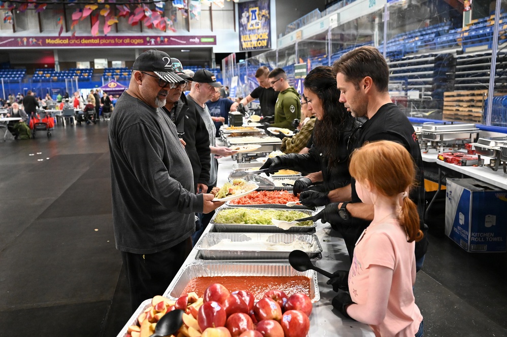 A community meal service event is taking place inside what appears to be an indoor arena or sports facility. Volunteers wearing black gloves are serving food from a long buffet-style table lined with aluminum trays containing various dishes. Attendees, including adults and a young child, are moving along the serving line to receive food.