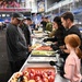 A community meal service event is taking place inside what appears to be an indoor arena or sports facility. Volunteers wearing black gloves are serving food from a long buffet-style table lined with aluminum trays containing various dishes. Attendees, including adults and a young child, are moving along the serving line to receive food.