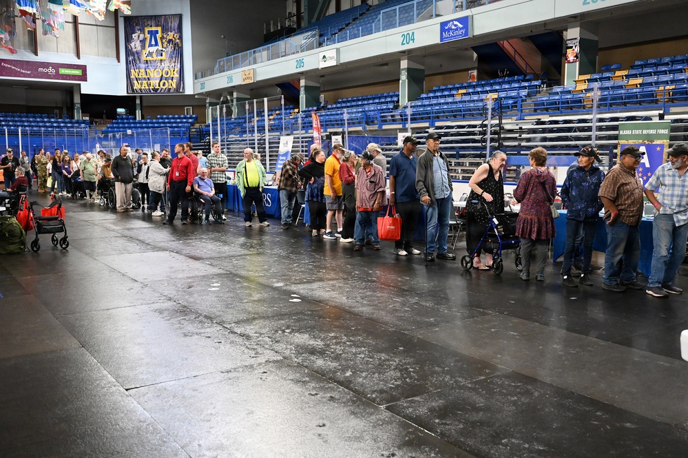 A large indoor arena, identifiable as the Carlson Center at the University of Alaska Fairbanks based on the 'Nanook Nation' banner, is hosting what appears to be a veterans' resource fair or stand-down event. A long line of attendees, including elderly individuals using walkers and wheelchairs, stretches across the arena floor past multiple vendor or service tables. A banner for the 'Alaska State Defense Force' is visible among the exhibitors.