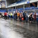 A large indoor arena, identifiable as the Carlson Center at the University of Alaska Fairbanks based on the 'Nanook Nation' banner, is hosting what appears to be a veterans' resource fair or stand-down event. A long line of attendees, including elderly individuals using walkers and wheelchairs, stretches across the arena floor past multiple vendor or service tables. A banner for the 'Alaska State Defense Force' is visible among the exhibitors.