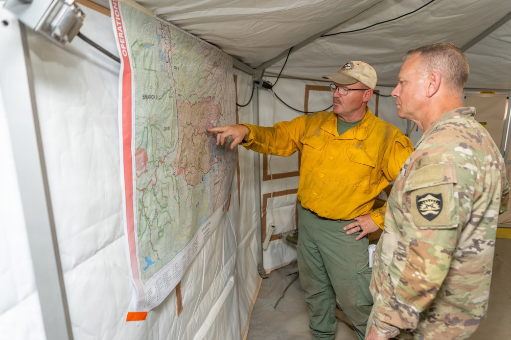 Two individuals are inside a field operations tent reviewing a large topographic map mounted on the wall. One person wearing a yellow wildfire firefighter shirt and tan cap is pointing to a specific location on the map, while a military personnel in OCP (Operational Camouflage Pattern) uniform observes. The setting appears to be an incident command or operations briefing environment.