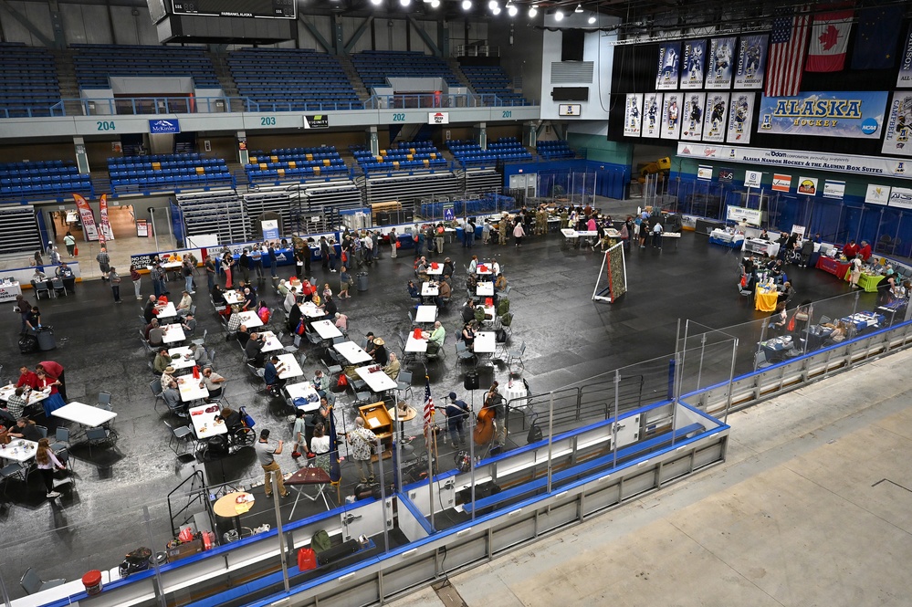 An indoor event is being held on the floor of an ice hockey arena in Fairbanks, Alaska, with the ice removed or covered. The venue appears to be hosting a community or career fair, with numerous tables, chairs, and attendees spread across the arena floor. Signage for 'Alaska Hockey' is prominently visible in the upper right corner of the arena.