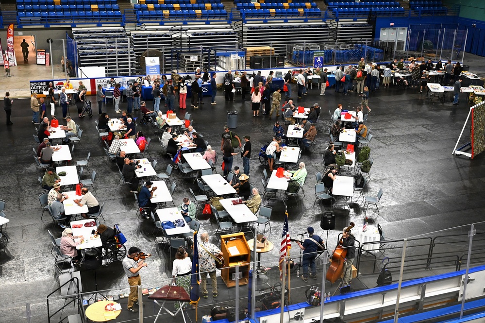 An aerial view of a large indoor arena or convention center hosting what appears to be a community event or veterans gathering, with numerous white rectangular tables arranged across the floor where attendees are seated and mingling. An American flag is visible near the bottom center, and musicians appear to be performing near the foreground. Vendor or information booths line the perimeter of the space.