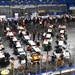 An aerial view of a large indoor arena or convention center hosting what appears to be a community event or veterans gathering, with numerous white rectangular tables arranged across the floor where attendees are seated and mingling. An American flag is visible near the bottom center, and musicians appear to be performing near the foreground. Vendor or information booths line the perimeter of the space.