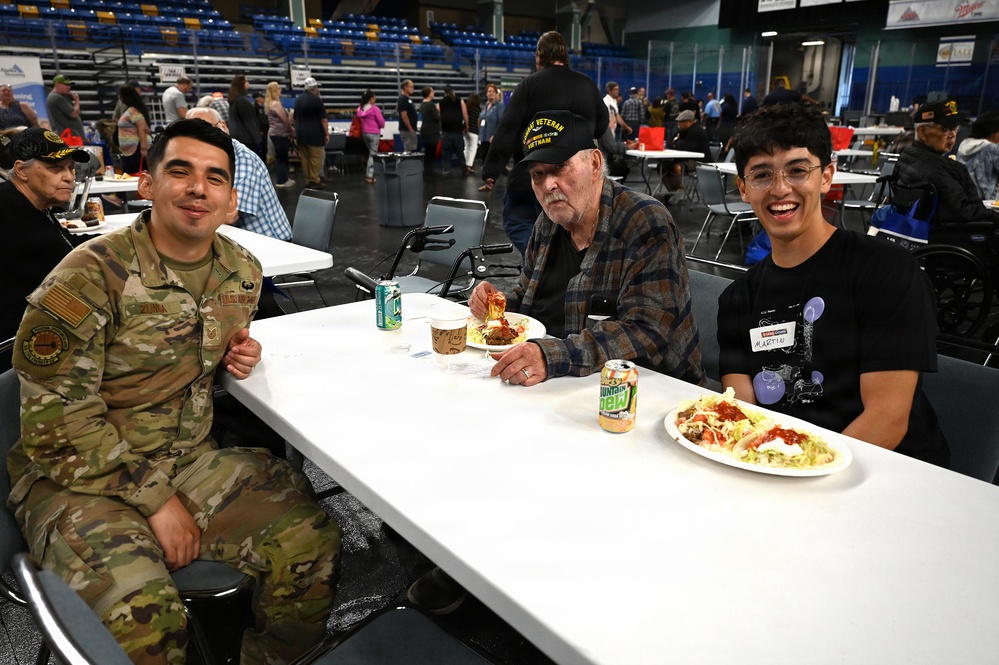 Three individuals are seated at a white table in a large indoor arena or convention center, sharing a meal together. The group includes a person in military camouflage uniform, an elderly man wearing a black 'Vietnam Veteran' cap, and a younger person wearing a name tag. The setting appears to be a veterans appreciation event or community meal gathering.