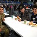 Three individuals are seated at a white table in a large indoor arena or convention center, sharing a meal together. The group includes a person in military camouflage uniform, an elderly man wearing a black 'Vietnam Veteran' cap, and a younger person wearing a name tag. The setting appears to be a veterans appreciation event or community meal gathering.