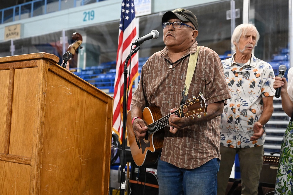Two older men are performing music at what appears to be an indoor arena or stadium venue. The primary performer in the foreground is playing an acoustic guitar and singing into a microphone at a podium, while a second man in a floral shirt holds a handheld microphone behind him. An American flag is visible in the background alongside stadium seating.