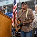 Two older men are performing music at what appears to be an indoor arena or stadium venue. The primary performer in the foreground is playing an acoustic guitar and singing into a microphone at a podium, while a second man in a floral shirt holds a handheld microphone behind him. An American flag is visible in the background alongside stadium seating.